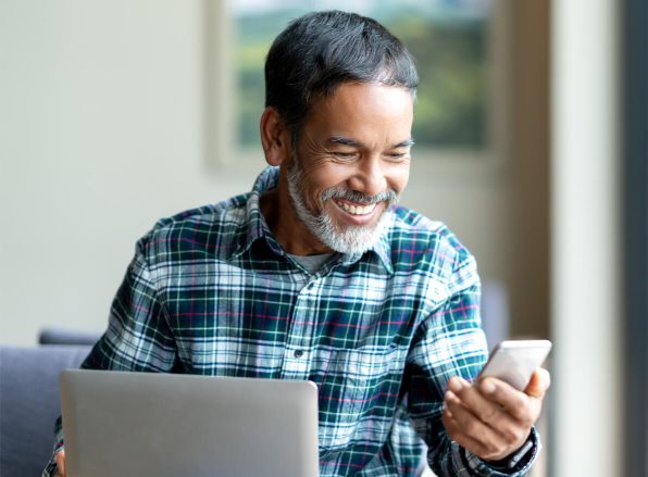 man smiling while looking at his mobile phone, with a laptop off to the side